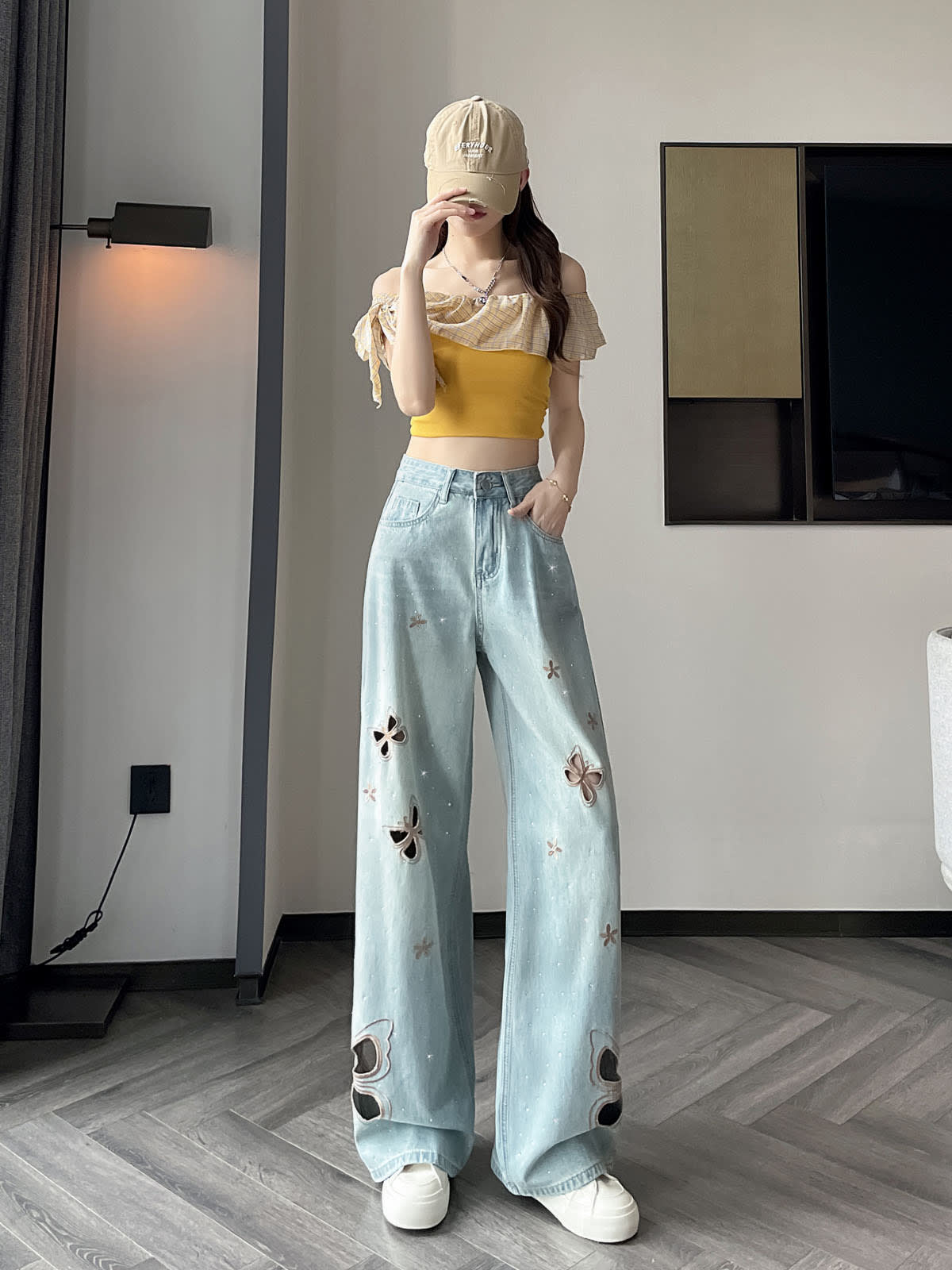 YEEYEEWang 라이트 블루 와이드레그 진, 아시안핏, 전신 스탠딩 포즈, 한 손은 주머니에 넣고 한 손은 캡 챙을 만지는 모습, 옐로우 러플 오프숄더 탑, 다리 라인의 나비 컷아웃과 플라워 스티치.