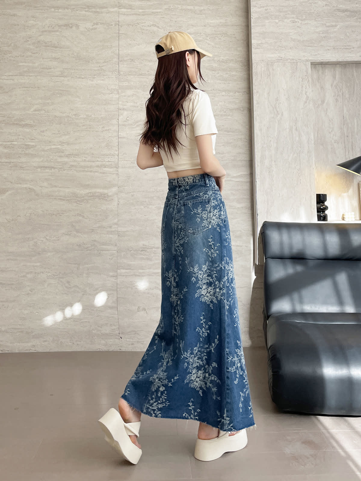 YEEYEEWang 블루 데님 맥시 스커트, 아시안핏, 뒷면 전신, 걷는 포즈, 베이지 캡, 뒷 패치 포켓과 움직이는 플레어 밑단의 플로럴 데님.