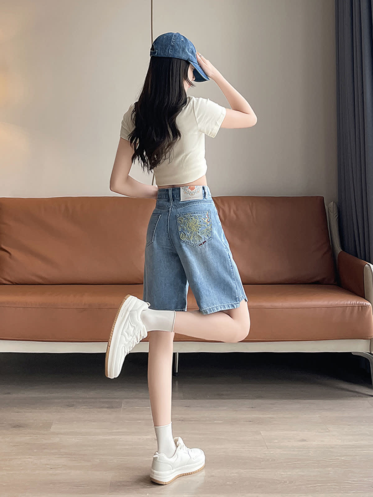 YEEYEEWang 블루 데님 버뮤다 쇼츠, 아시안핏, 전신 뒤쪽 3/4 각도, 한 다리를 뒤로 든 포즈, 손으로 캡 챙을 만지며 뒷주머니 자수가 보임.